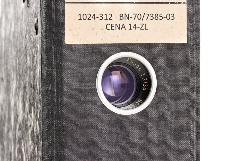 Close-up of a camera lens labeled "Xenon 1:2/35" mounted on a textured black surface, next to a paper label with codes "1024-312 BN-70/7385-03 CENA 14-ZL".