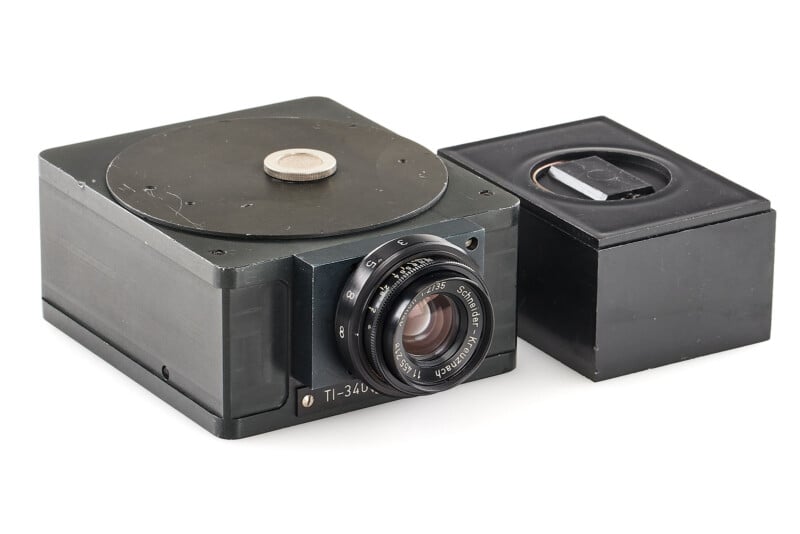 A vintage black camera with a lens on the front sits next to a matching square black accessory, both on a white background.