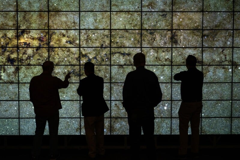 Four people stand silhouetted in front of a large, brightly lit screen displaying a detailed image of a star-filled section of space or a galaxy.