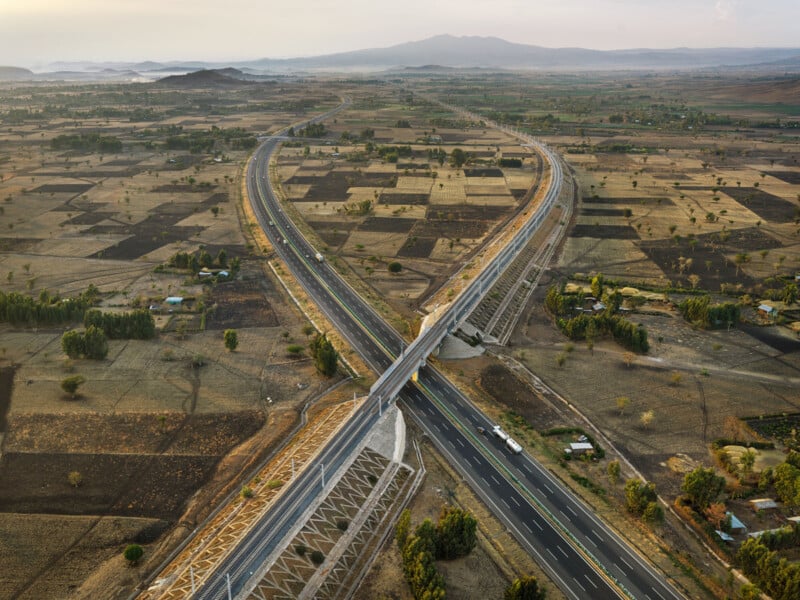 Aerial view of two intersecting highways forming an X shape, surrounded by dry, patchwork farmland with scattered trees and distant mountains under a hazy sky.