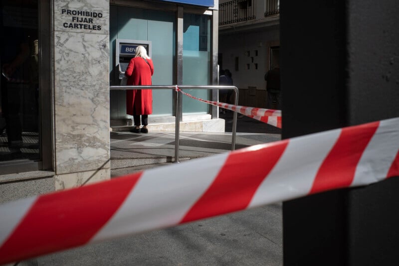 A person in a red coat and white headscarf uses an outdoor ATM. Red and white caution tape blocks access, and a sign on the wall reads "PROHIBIDO FIJAR CARTELES.