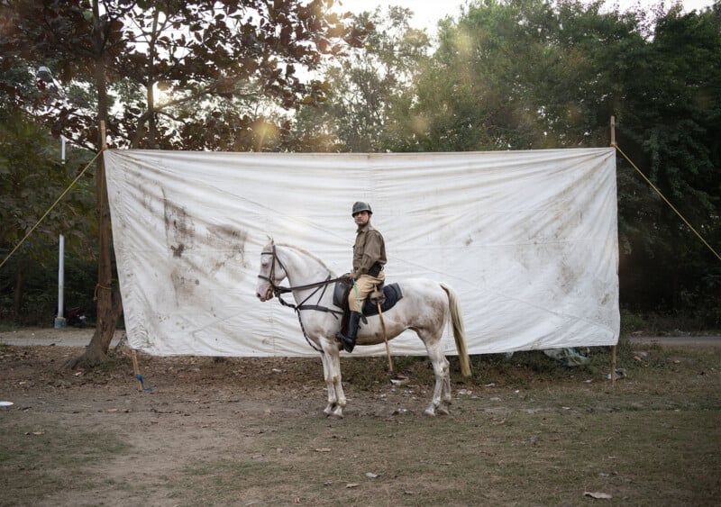 A person in a uniform and helmet sits on a white horse in front of a large, dirty white sheet backdrop outdoors, with trees and sunlight in the background.
