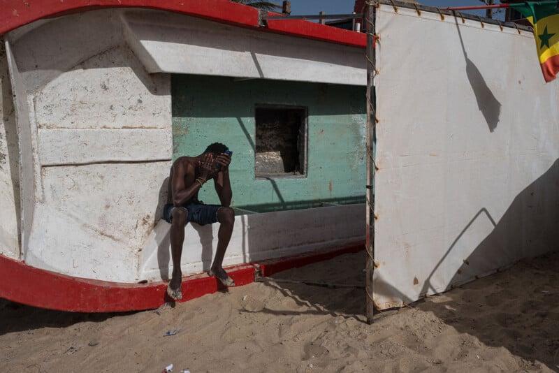 A man sits on a beached, overturned boat, covering his face with his hands. He is in the shade, next to a white partition with the Senegalese flag, on a sandy beach under a clear sky.