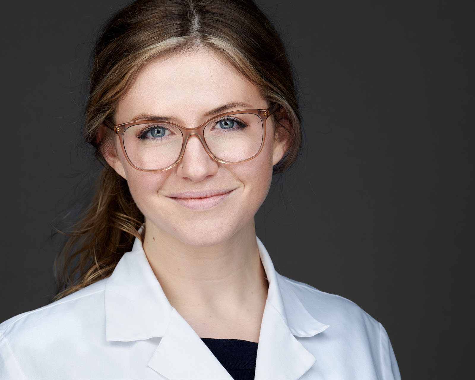 A young woman with light brown hair, wearing glasses and a white lab coat, smiles confidently at the camera against a dark gray background.