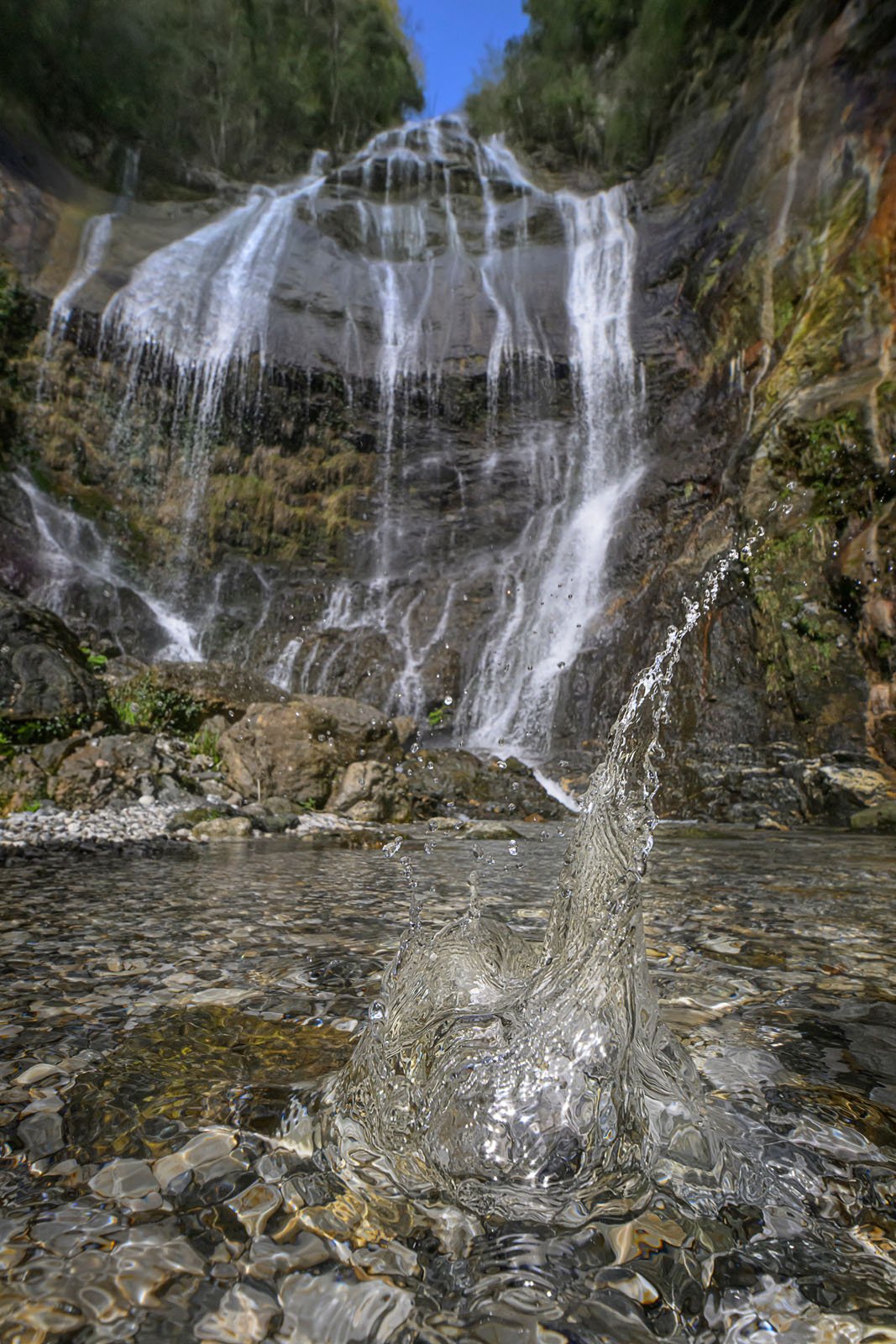 Clear water splashes upward in the foreground, creating a splash crown effect, with a tall, cascading waterfall flowing over rocky cliffs and lush greenery in the background under a blue sky.