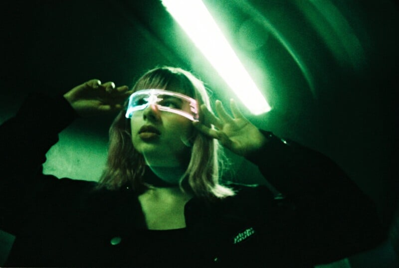 A person wearing futuristic, glowing neon glasses stands under a green light, with hands raised near their face, creating a sci-fi, cyberpunk atmosphere. The background is dark and atmospheric.
