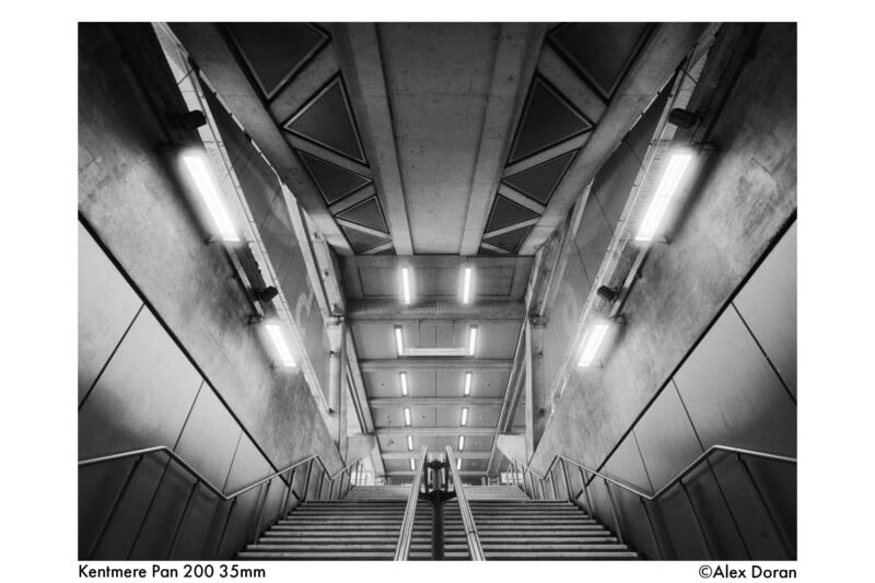 Black and white photo of a modern staircase in a concrete building, taken from the bottom looking up. Fluorescent lights and geometric ceiling patterns are visible, with handrails leading up the stairs.