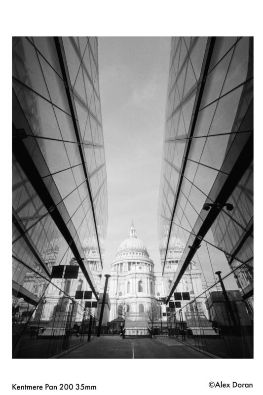 Black-and-white photo of St. Paul’s Cathedral framed symmetrically by two modern glass buildings, reflecting the cathedral’s dome. Taken from a low angle, creating a dramatic, geometric perspective.