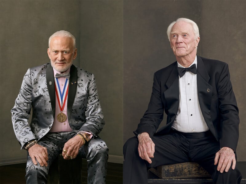 Two older men in formal attire sit against a neutral backdrop. The man on the left wears a silver patterned suit with medals and jewelry. The man on the right wears a classic black tuxedo. Both look slightly off camera.