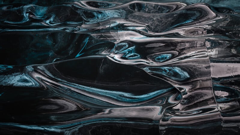 Close-up of abstract, wavy patterns and textures in a dark, translucent material, possibly ice or glass, with reflective surfaces and hints of blue and silver tones.