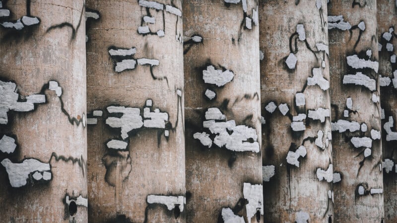 Vertical concrete columns with irregular white patches and dark stains, creating a weathered, abstract pattern. The surface texture is rough, and the columns are closely aligned side by side.