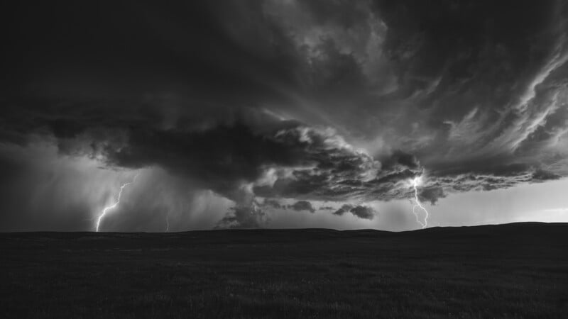A dramatic black and white photo of a stormy sky over open grassland, with dark, swirling clouds and two bright lightning bolts striking the ground in the distance.