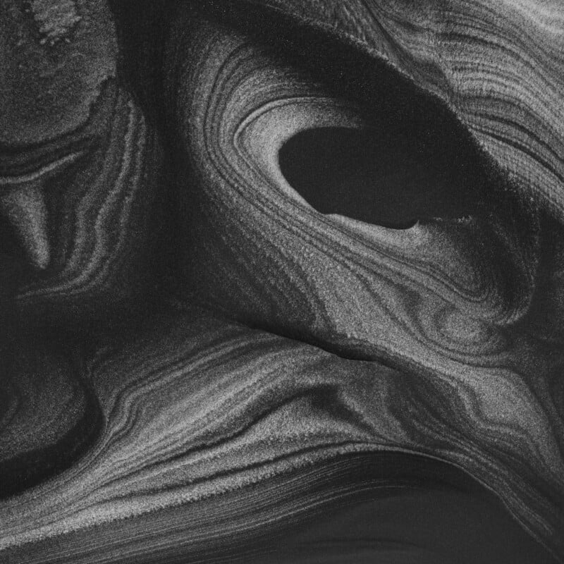 Abstract black and white image showing swirling, layered patterns resembling marbled sand or stone, with varying shades of gray creating a sense of depth and texture.