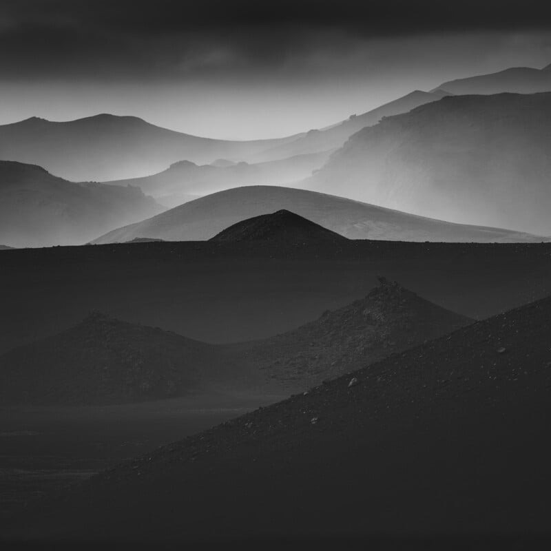 Black and white image of rolling hills and mountains, layered in mist. The foreground features dark, rocky terrain, while the background shows lighter, softer silhouettes of hills receding into the distance.