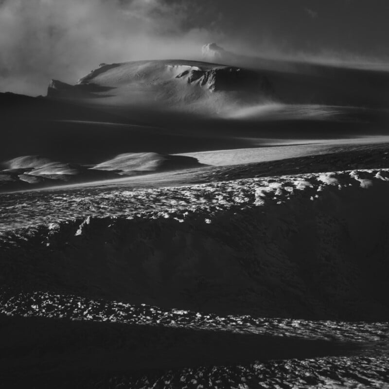 A dramatic black and white photo of a snow-covered mountain ridge, with light and clouds creating striking shadows and highlights across the rugged landscape.