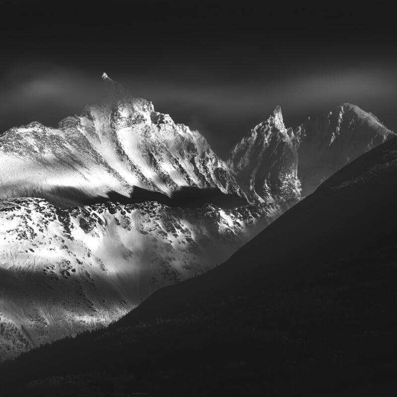 Black and white photo of dramatic, snow-covered mountain peaks with sharp ridges, illuminated by sunlight against a dark sky, creating striking contrasts and deep shadows on the landscape.
