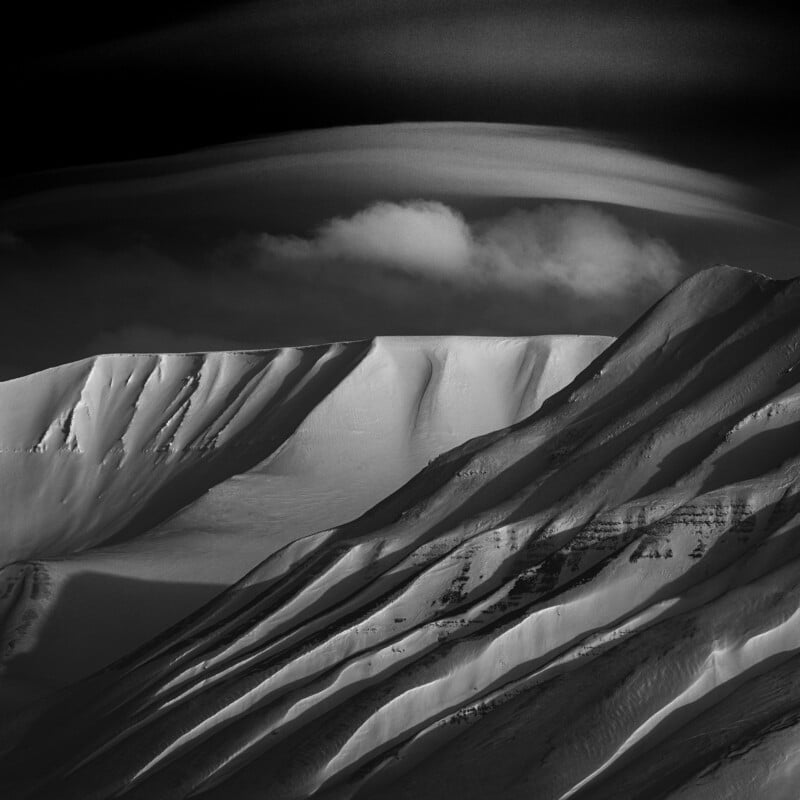 Black and white photo of snow-covered mountain slopes with dramatic ridges and shadows, beneath a dark sky with a single white cloud and streaks of light.