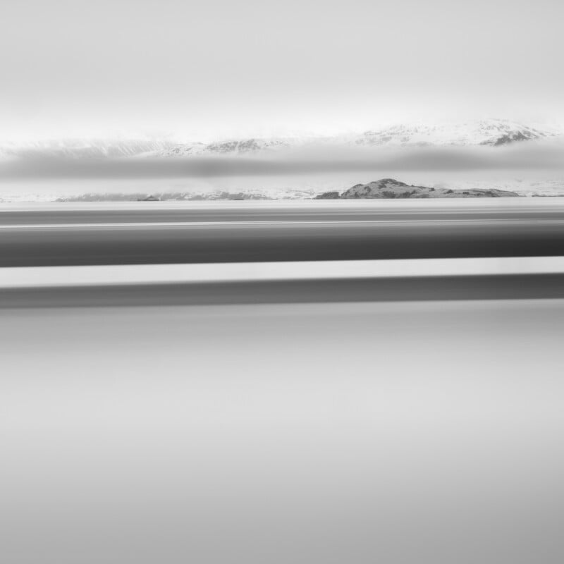 Minimalist black-and-white landscape with smooth, blurred horizontal lines suggesting water or road in the foreground, a distant rocky hill, and snow-covered mountains beneath a cloudy sky in the background.