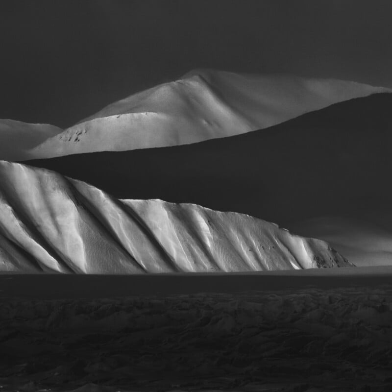 Snow-covered mountains and ridges cast dramatic shadows under low light, creating a stark contrast between illuminated and dark areas in a rugged, wintry landscape.