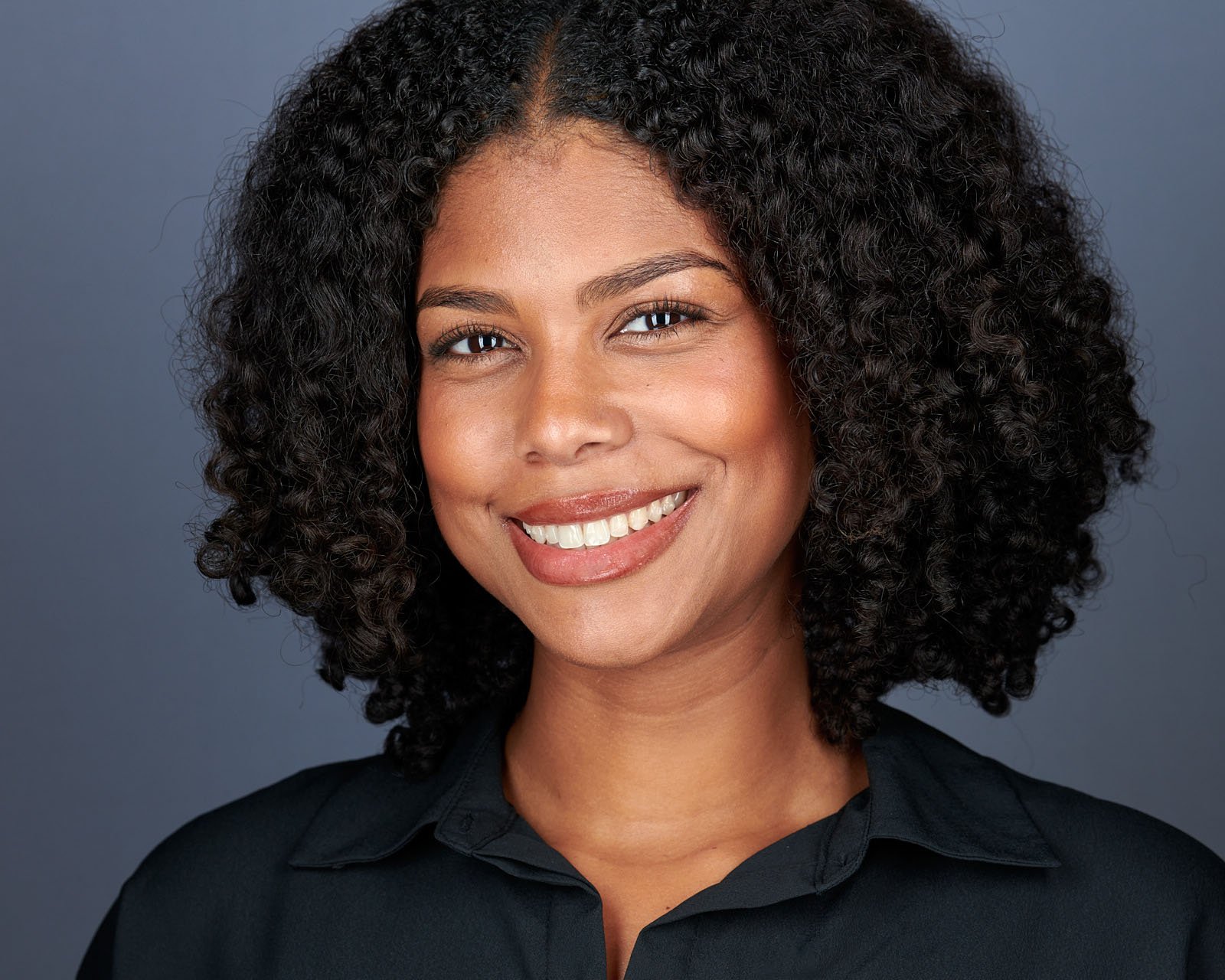 A woman with curly dark hair and medium skin tone smiles confidently. She is wearing a black collared shirt and is posed against a plain, dark gray background.