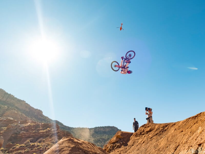 A mountain biker performs a backflip high in the air off a dirt ramp, while a photographer captures the moment below under a bright blue sky with the sun shining.