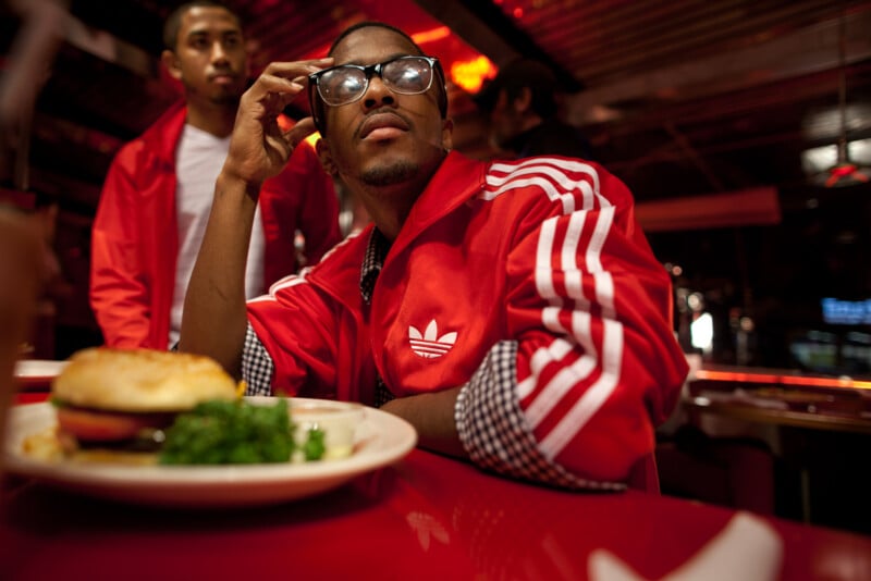A man wearing glasses and a red Adidas jacket sits at a red counter, resting his head on his hand, with a plate of food in front of him. Another person in a similar red jacket stands in the background.