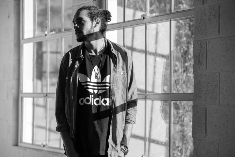 A man with a beard and tied-back hair stands by large windows, wearing a dark Adidas shirt and an unbuttoned shirt over it. Sunlight streams in, casting shadows on the wall and floor. The image is in black and white.