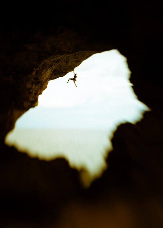 A rock climber hangs from the edge of a cliff, silhouetted against a bright sky, as seen through a cave opening with blurred, dark edges framing the scene.