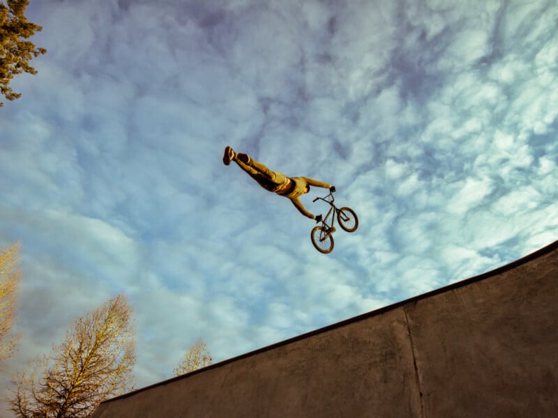 A person performs a dramatic stunt on a BMX bike, leaping off a ramp with their body fully extended in midair against a cloudy sky, above a concrete skate park and bare trees.