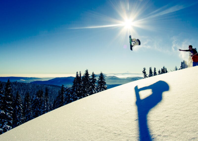A snowboarder catches air high above a snowy slope, with their shadow cast on the snow. The sun shines brightly in a clear blue sky, while pine trees and distant mountains are visible in the background.