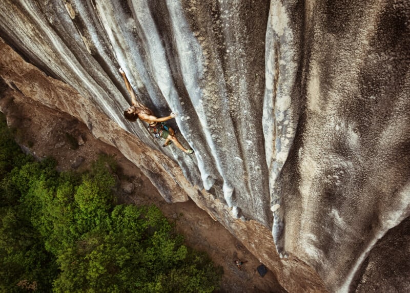 A rock climber ascends a steep, textured cliff face, reaching up with one arm. The ground and green trees are far below, emphasizing the height and challenge of the climb.