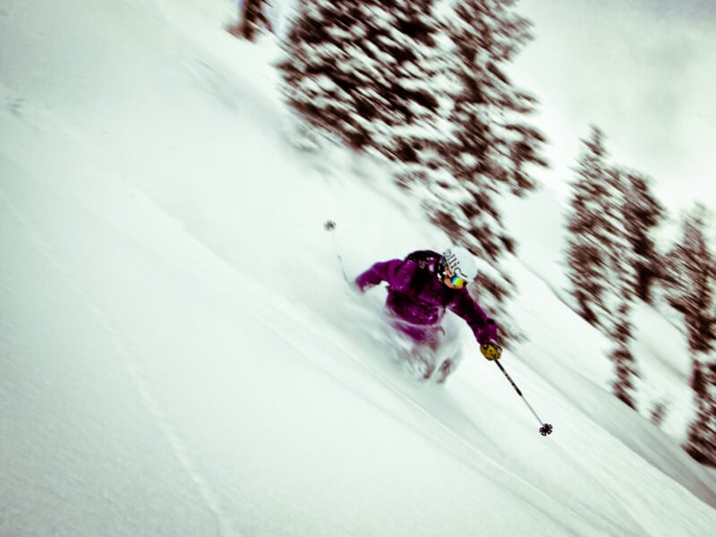 A skier in a purple jacket and helmet glides quickly down a snowy slope, surrounded by snow-covered trees. The skier is using poles and carving through fresh powder under a cloudy sky.