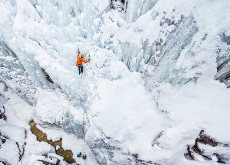 An ice climber wearing an orange jacket ascends a steep, icy wall surrounded by snow and frozen formations, viewed from above in a dramatic winter landscape.