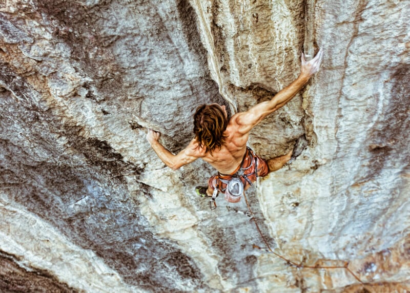 A rock climber with long hair and no shirt ascends a steep, rugged cliff, reaching out with chalked hands and wearing a harness with climbing gear attached.