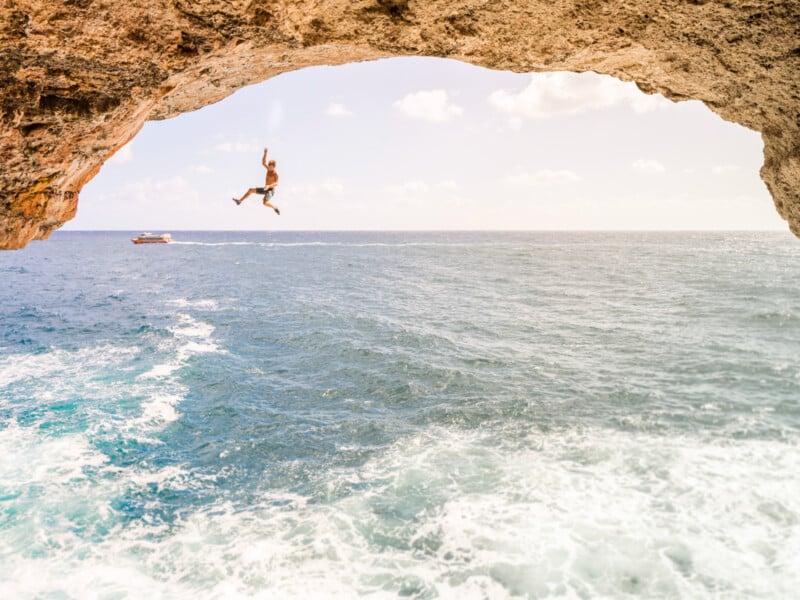 A person is captured mid-air after jumping off a rocky cliff over the ocean, with waves below and a boat in the distance under a partly cloudy sky.