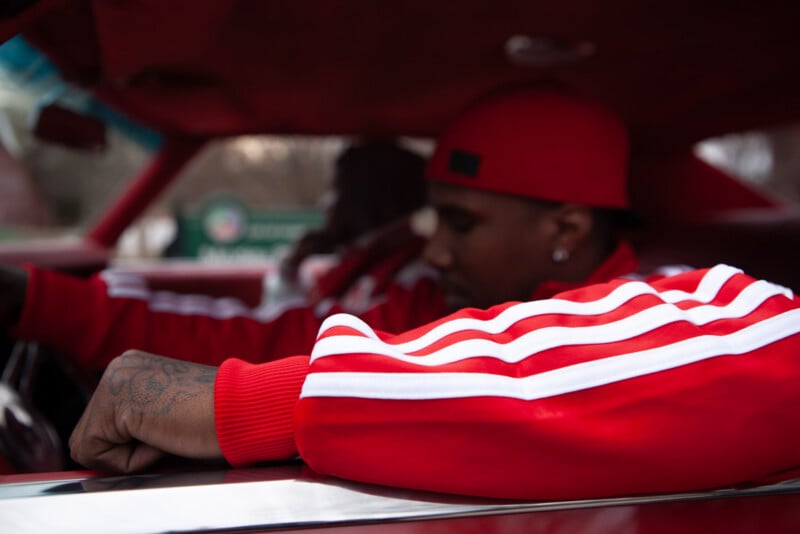Two people sit inside a car, both wearing matching red and white striped jackets. The person in the foreground rests their arm on the car door, while the driver appears focused ahead. The interior is dimly lit.
