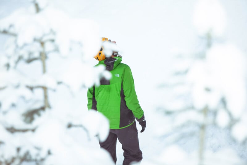 A person wearing a bright green jacket and orange helmet stands outdoors in a snowy landscape, partially obscured by snow-covered tree branches in the foreground.