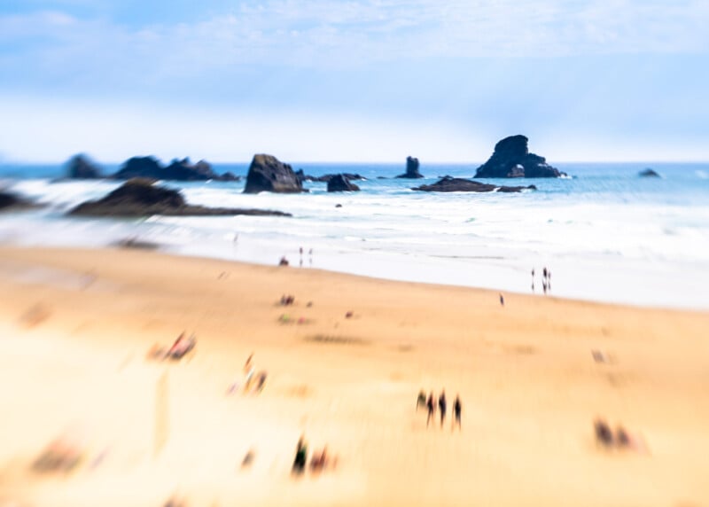 A sandy beach with scattered people relaxing and walking, with large rocks in the ocean and waves crashing under a mostly cloudy sky. The image is slightly blurred, creating a dreamy effect.