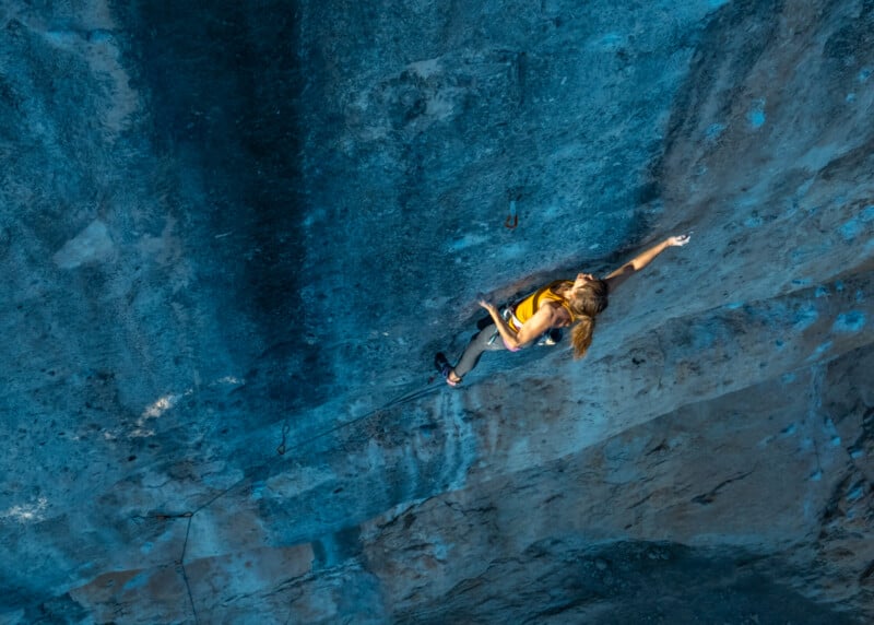 A climber in a yellow tank top ascends a steep, blue-toned rock wall, using both hands for balance and looking upward, with safety gear attached to the wall above.