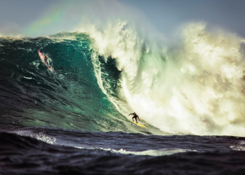 Two surfers ride a massive ocean wave; one is near the wave’s peak while the other maneuvers down the face, surrounded by crashing whitewater and dramatic spray.