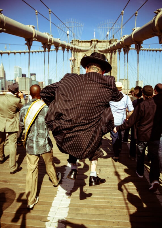 A person in a pinstripe suit and hat jumps energetically on the Brooklyn Bridge among a crowd of people, with the city skyline visible in the background on a sunny day.