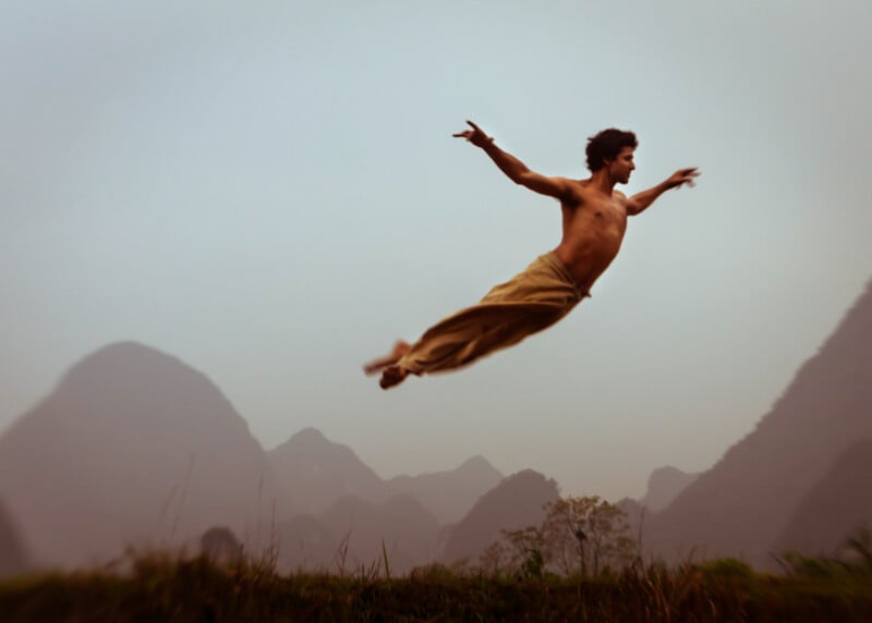 A shirtless man wearing loose pants leaps horizontally in mid-air against a misty backdrop of mountains and grass, arms outstretched, appearing to fly or dance energetically.