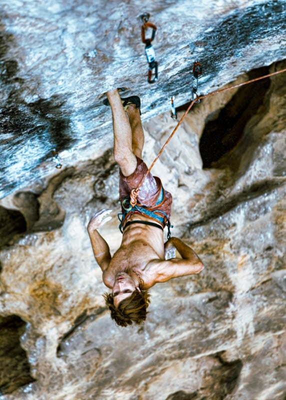 A shirtless rock climber hangs upside down by his legs on a steep, rocky overhang, gripping the rock with chalked hands and secured by safety ropes and harness. The rugged cliff face fills the background.
