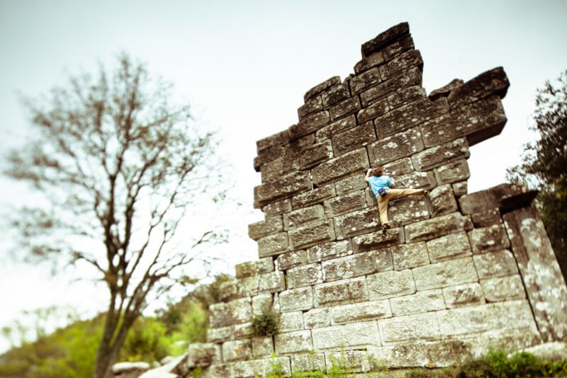 A person wearing a blue shirt and beige pants climbs a large, weathered stone wall outdoors with a tree in the background and cloudy sky above.