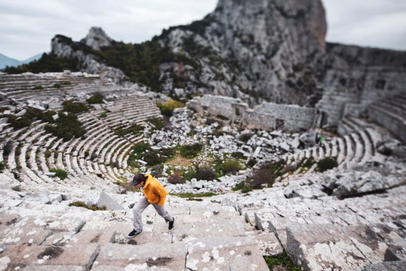 A person in a yellow jacket walks up stone steps in an ancient, ruined amphitheater surrounded by rocky mountains and greenery. The scene is blurred except for the person in focus.