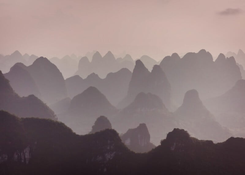 Misty, overlapping karst mountains with rounded peaks fade into the distance under a soft, muted pink sky, creating a dreamy and tranquil landscape.