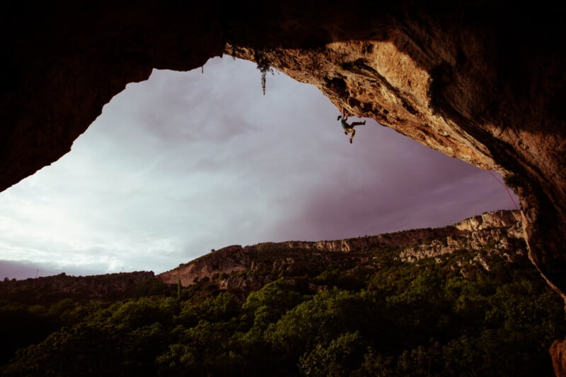 A rock climber hangs from the underside of a large overhanging cliff, silhouetted against a cloudy sky, with green trees and rocky hills visible below in the distance.