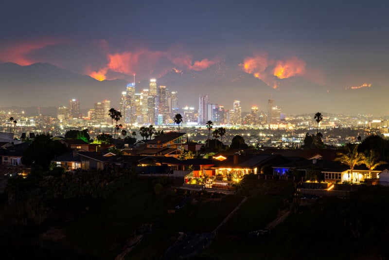 A nighttime cityscape of Los Angeles with illuminated buildings in the foreground and wildfires burning on the hills in the background, casting an orange glow and smoke into the night sky.