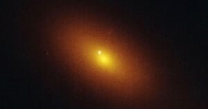 A bright yellow-orange glowing spot surrounded by a hazy, diffuse halo set against a dark background, resembling a distant galaxy or luminous celestial object in space.