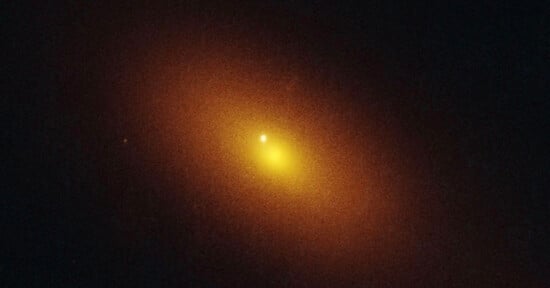 A bright yellow-orange glowing spot surrounded by a hazy, diffuse halo set against a dark background, resembling a distant galaxy or luminous celestial object in space.